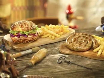 A burger with fries and a grilled spiral sausage with fries served on wooden boards at Au Chalet de la Marionnette in Disneyland Park, with a Jiminy Cricket figurine and a cuckoo clock in the Pinocchio-themed background