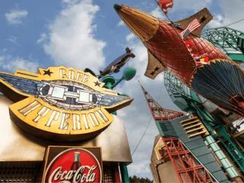 The dramatic exterior of Café Hyperion at Disneyland Park, featuring a bold aviation-themed sign, a giant airship and colourful flags against a bright blue sky in Discoveryland