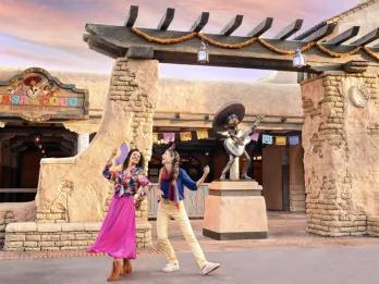 Two guests dancing outside Casa de Coco Restaurante de Familia at Disneyland Park, with a Miguel statue, colourful papel picado bunting and a vibrant Mexican-inspired entrance at sunset