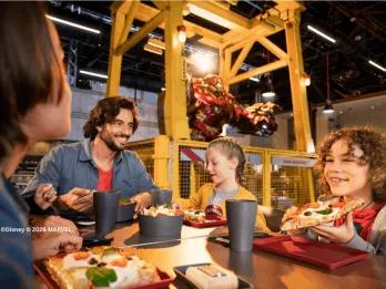 A family enjoying pizza and salads at Stark Factory in Disney Adventure World, smiling together in an industrial Marvel-themed setting with a Hulkbuster Iron Man suit suspended in the background