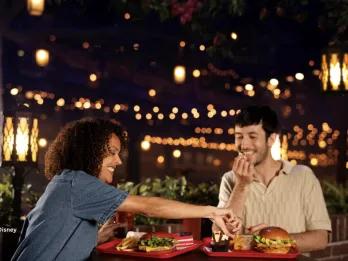 A couple sharing burgers, fries and desserts at The Hollywood Gardens Restaurant at Disney Adventure World, laughing together under warm string lights and floral canopy in a glamorous outdoor evening setting