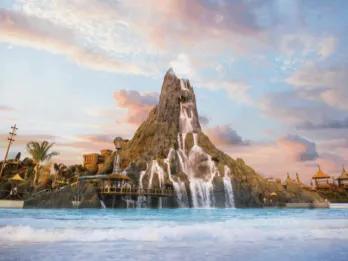 A wide shot of a massive artificial volcano with cascading waterfalls flowing into a large, wavy blue pool, under a pastel pink and blue sunset sky with scattered clouds