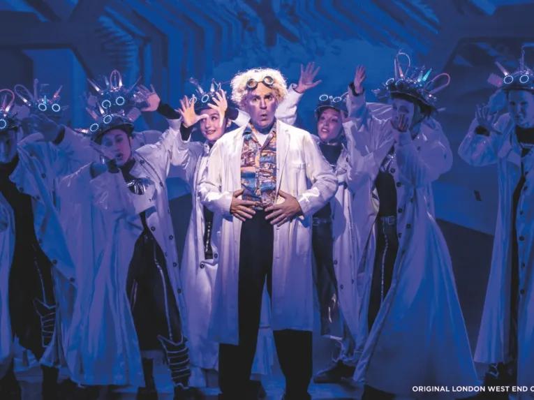 Doc Brown wearing a white lab coat standing centre stage in front of dancers also wearing lab coats