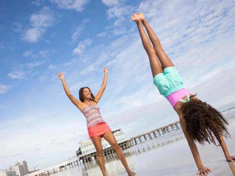 A mother cheering while her daughter does a handstand on the sand near the Daytona Beach Pier