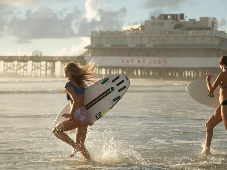 Two surfers carrying surfboards and running into the ocean near the Daytona Beach Pier