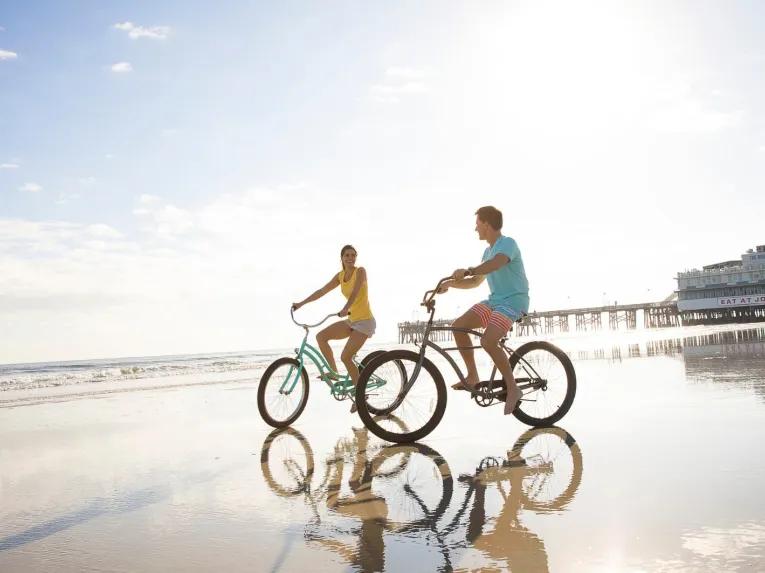 A couple riding cruiser bicycles along the hard-packed sand of Daytona Beach next to the ocean.