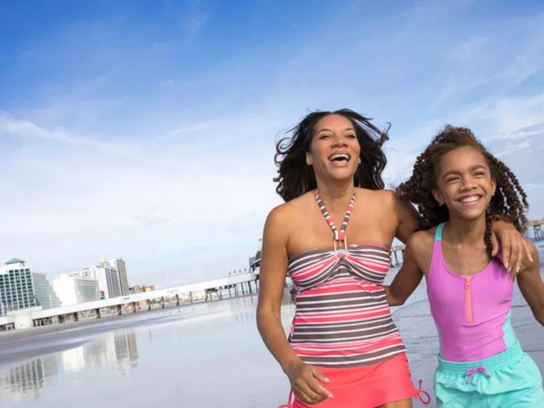 A happy mother and daughter laughing on Daytona Beach with the pier and hotels in the background.
