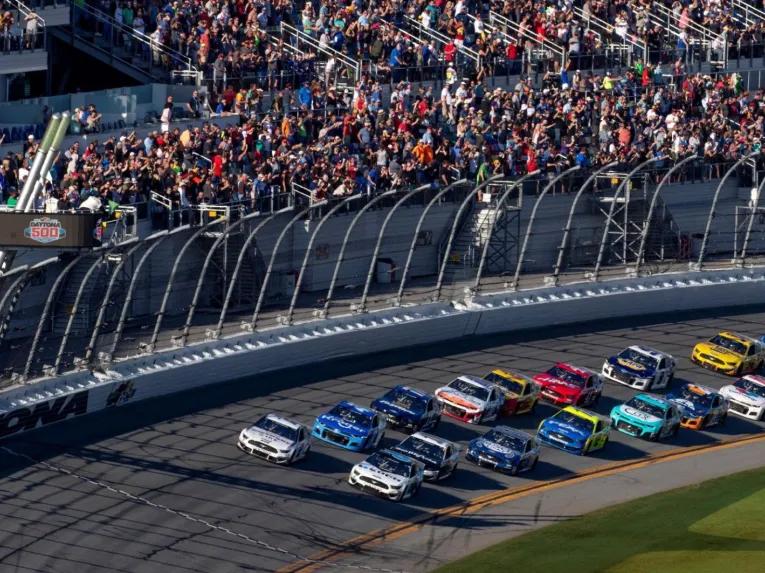 NASCAR race cars speeding past packed grandstands during a race at the Daytona International Speedway.